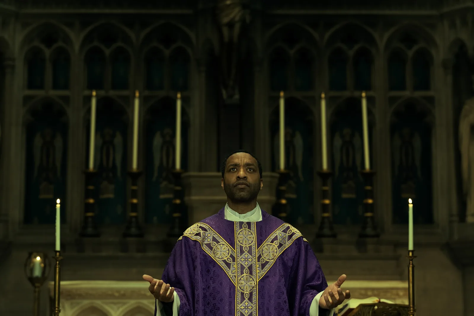 Chiwetel Ejiofor en prêtre, chasuble violette, les bras ouverts devant un autel aux hauts cierges, dans The Exorcist: Martyrs